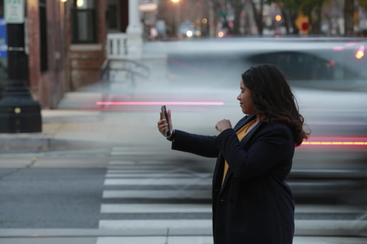 A dark-skinned female wearing a black jacket holding her phone in her right hand. She is looking at her phone and signing with her left hand in front of a busy road behind her.